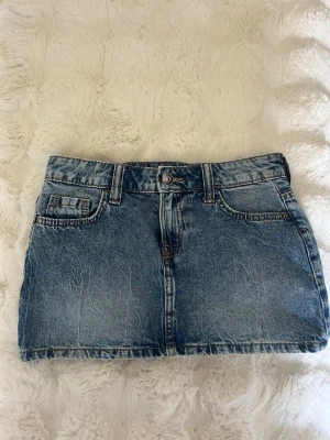 Blå jeanskjol med fickor - Snygg blå jeanskjol i klassisk modell med fem fickor, bälteshällor och knappstängning framtill. Kjolen har en kort passform och är tillverkad i denim med slitna detaljer för en cool look. Perfekt att matcha med en t-shirt eller hoodie. Storlek xs. 