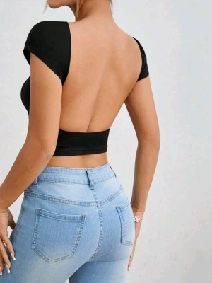 Svart croppad topp med öppen rygg - Snygg svart croppad topp med korta ärmar och helt öppen rygg. Toppens tighta passform framhäver figuren och den är tillverkad i ett stretchigt material som känns bekvämt mot huden. Perfekt för dig som vill ha en stilren men ändå vågad look.