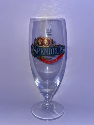 Spendrups ölglas med logga - Klassiskt ölglas från Spendrups med färgglad logga framtill. Glaset är genomskinligt och har en rundad form med fot. Perfekt för att servera öl eller andra drycker. Rymmer 0,5 liter och har tydlig märkning upptill.
