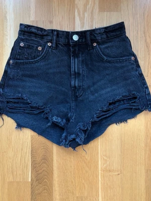 Svarta high waist jeansshorts - Säljer ett par svarta högmidjade jeansshorts med råa, slitna kanter och fransar. Shortsen har fem fickor. Perfekta för en avslappnad och trendig look.
