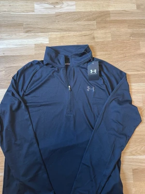 Svart träningströja från Under Armour - Svart långärmad träningströja från Under Armour med halv dragkedja och hög krage. Tillverkad i ett lätt och stretchigt funktionsmaterial som andas. Perfekt för löpning, gym eller annan träning. Diskret logga på bröstet. Skick 10/10 aldrig använd.