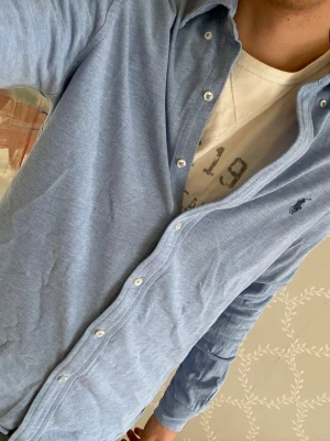 Ljusblå Ralph Lauren skjorta - Snygg Ralph Lauren i modellen Featherweight Mesh. Skjortan har klassisk krage, knappar framtill och broderad logga på bröstet. Tillverkad i mjuk bomull som känns lätt och bekväm. Perfekt för en stilren look.