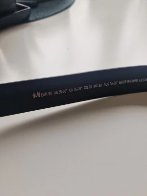 Svart bälte från H&M i skinnimitation - Stilrent svart bälte från H&M i skinnimitation med klassiskt spänne i svart metall. Perfekt accessoar för att lyfta din outfit och passar till både jeans och kostymbyxor. Bältet har flera hål för justerbar passform.