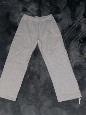 Grå baggy mjukisbyxor från Nike - Gråa mjukisbyxor från Nike med rak passform och resår i midjan samt dragsko. Byxorna har sidofickor och en liten vit Nike-logga broderad på ena benet. Perfekta för chill dagar eller träning. Aldrig använda helt nya!