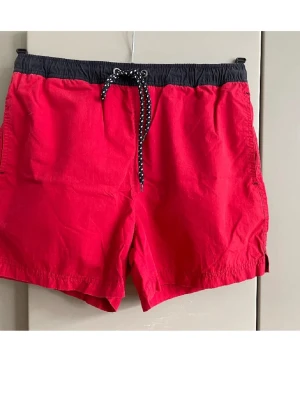 Röda badbyxor med svart midja - Snygga röda badbyxor med svart resårmidja och vitprickigt snöre. Klassisk modell med sidfickor och en bakficka med etikett. Perfekta för stranden eller poolen. Materialet känns lätt och snabbtorkande.