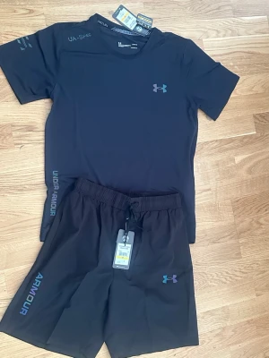 Svart träningsset från Under Armour - Snyggt svart träningsset från Under Armour med t-shirt och shorts. Båda plaggen har logga och tryck i blått och lila. T-shirten är kortärmad och shortsen har resår i midjan samt dragsko. Perfekt för gym eller löpning.