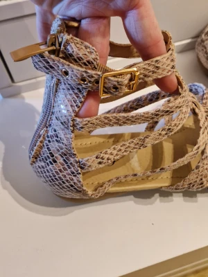 Beige sandaler med ormskinnsmönster - Snygga sandaler i beige med ormskinnsmönster och flera smala remmar över foten. Sandalerna har en öppen tå och justerbart spänne i guld vid ankeln. Materialet är konstläder med glansig finish och sandalerna har platt sula.