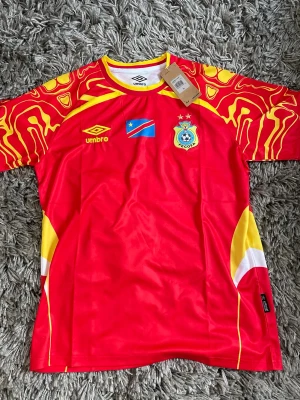 Röd FECOFA fotbollströja från Umbro - Snygg röd fotbollströja från Umbro med FECOFA-logga och Kongo-Kinshasas flagga på bröstet. Tröjan har gula och vita detaljer samt unikt mönster på ärmarna. Perfekt för dig som vill sticka ut på planen eller visa support för landslaget.
