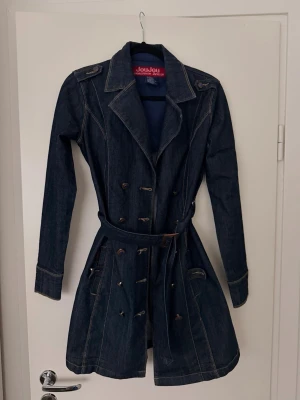 Mörkblå denim trenchcoat från Joujou - Sitter som storlek S!! Snygg mörkblå trenchcoat i denim från JoliYou med dubbelknäppning, bälte i midjan och klassisk krage. Jackan har långa ärmar, markerade sömmar och axelklaffar för en cool look. Perfekt för dig som gillar jeansstil och vill sticka ut. Funkar även bra som jeansklänning! 