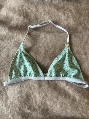 Mintgrön blommig bikiniöverdel - Söt mintgrön bikiniöverdel med små blommor och vita band. Triangelmodell med knyt i nacken och en liten snäcka som detalj på ena axelbandet. Perfekt för strandhäng och soliga dagar.