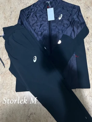 Svart träningsset från Asics - Snyggt svart träningsset från Asics med jacka och byxor. Jackan har dragkedja och diskret mönster på framsidan, samt Asics-logga på bröstet. Byxorna har resår i midjan och logga på benet. Perfekt för löpning eller gym.