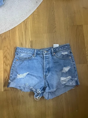 Blå slitna jeansshorts - Säljer ett par blå denimshorts med slitna detaljer och fransig kant. Klassisk femficksmodell med hög midja och knappgylf. Perfekta för varma dagar och en avslappnad stil.