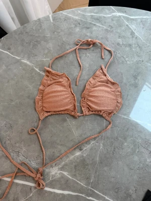 Glittrig rosa bikiniöverdel med volang - Bikiniöverdel i glittrigt rosa material med volangdetaljer längs kanterna.