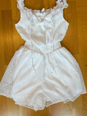 Vit kort jumpsuit från FB Sister/New Yorker  - Supersöt vit kort jumpsuit från FB Sister med volangkant vid axlarna, smock i midjan och små broderade prickar i tyget. Axelbanden är justerbara med knytband och materialet känns lätt och luftigt, perfekt för varma dagar. Katt finns i hemmet, skriv om ni har frågor. 