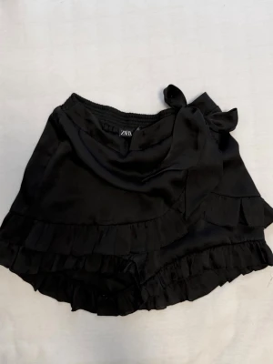 Svarta omlottshorts med volang Zara - Svarta omlottshorts från Zara i glansigt material med volangdetaljer längs kanterna och en söt knytning i midjan. Perfekta för en trendig och bekväm sommarlook.