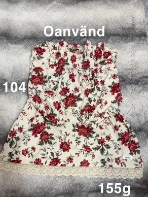 Blommig kortklänning från Newbie - Söt kortklänning från Newbie i vit bomull med röda och gröna blommor. Klänningen har långärmade ärmar och spetskant längst ner för en romantisk touch.🌸 Oanvänd. Storlek 104