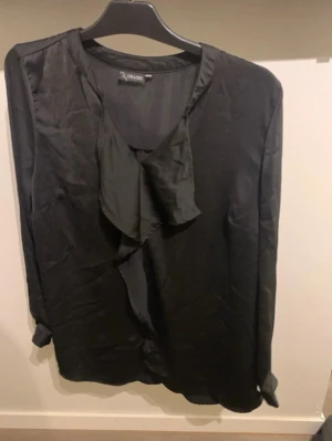 Svart blus med volangdetalj - Stilren svart blus från Cellbes med lång ärm och snygg volangdetalj framtill. Blusen har en mjuk, glansig finish och rund halsringning. Perfekt för dig som gillar en enkel men ändå chic look.