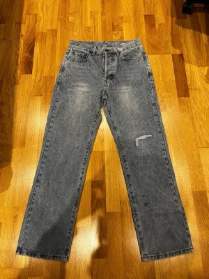Blå raka jeans från Levi's - Ett par grå raka jeans från Levi's med klassisk femficksdesign och vintagekänsla. Jeansen har en liten sliten detalj på ena benet och är tillverkade i kraftig denim. Perfekta för en avslappnad och trendig look.