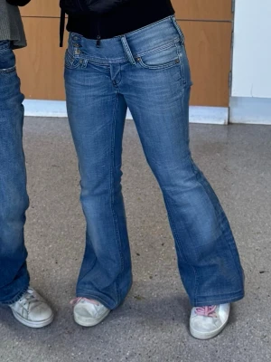 Blå bootcut jeans med låg midja - Säljer ett par klassiska blå bootcut jeans med låg midja och fem fickor. Jeansen har en snygg tvättad look och utsvängda ben. Perfekta för dig som gillar retrostil och vill ha ett par jeans som sticker ut lite extra.