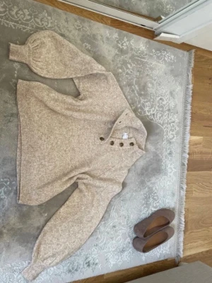 Beige stickad tröja med knappar - Mysig beige stickad tröja med breda ärmar och ribbade muddar. Tröjan har en hög krage med fyra dekorativa knappar framtill och är gjord i ett mjukt, melerat material. Perfekt för lager på lager och en avslappnad stil.