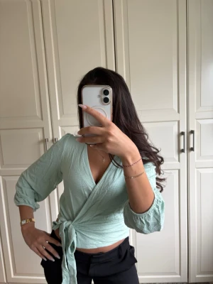 Ljusgrön omlottopp med knyt - Snygg ljusgrön omlottopp med knyt i midjan och trekvartsärmar. Toppen har v-ringning och är gjord i ett mjukt, lätt material som passar perfekt till både jeans och kjol. Enkel men trendig design som ger en fräsch look.