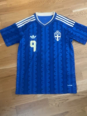 Blå Sverige fotbollströja nr 9 - Säljer en blå fotbollströja med Sveriges landslagsemblem och nummer 9 på bröstet och ryggen. Tröjan har vita detaljer, Adidas-logga och texten 'ISAK' på ryggen. Tillverkad i ett lätt funktionsmaterial med mönstrad design och vita ränder på axlarna.