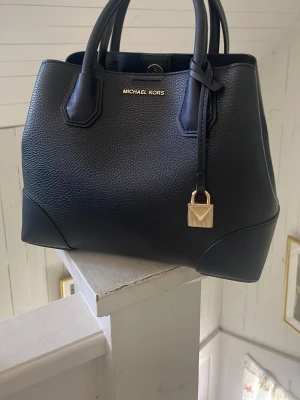 Svart handväska från Michael Kors - Stilren svart handväska från Michael Kors i kraftigt läder med guldfärgade detaljer och dekorativt hänglås. Väskan har både handtag och en avtagbar axelrem för flexibel användning. Klassisk fyrkantig form och diskret logga framtill.