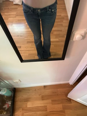 Blå bootcut jeans från True Religion - Använt 4 gånger!! Säljer ett par blå bootcut jeans från True Religion med klassiska kontrastsömmar och detaljerade bakfickor. Jeansen har låg midja och stängs med dragkedja och knapp. Perfekta för en avslappnad och trendig look. 