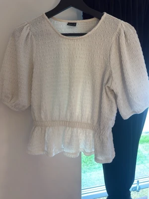 Vit smockad blus med puffärm - Söt vit blus med smockad struktur och puffiga korta ärmar. Blusen har rund halsringning och markerad midja med volang nedtill. Tillverkad i ett lätt och luftigt tyg som ger en romantisk känsla.