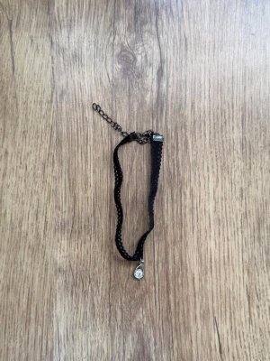 Svart choker med droppformad pärla - Snygg svart choker i spetsliknande band med en liten droppformad pärla som hänge. Halsbandet har justerbar kedja i metall och stängs med ett klassiskt spänne. Perfekt för att ge din outfit en edgy och trendig touch.