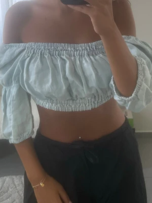 Ljusblå offshoulder croppad topp - Säljer en ljusblå croppad topp med offshoulder och puffiga trekvartsärmar. Toppen har resår vid axlar och midja för en snygg passform. Perfekt för varma dagar och ger en trendig vibe till din outfit.
