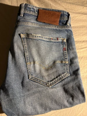 Ljusblå slim fit jeans Replay - Säljer ett par ljusblå slim fit jeans från Replay med klassisk femficksdesign och snygga slitningar. Jeansen har en skön passform och detaljer som Replay-lappen bak i midjan och diskreta röda sömmar vid fickan. Perfekta för en avslappnad och trendig look. Dem är sydda inuti och under som man ser på bilden. Lite utgivna vid fickorna och längs ner vid byxan. Inga stora fläckar eller skador.