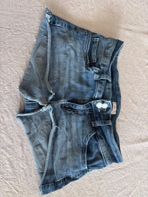 Blå jeansshorts från Nelly - Säljer ett par blå jeansshorts från Nelly med råa kanter och slitningar. Shortsen har klassisk femficksdesign, två knappar framtill och bälteshällor. Perfekta för varma dagar och en avslappnad stil.