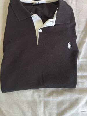 Svart pikétröja med vit logga - Klassisk svart pikétröja med vit knappslå och broderad logga på bröstet. Tröjan har korta ärmar och krage, perfekt för en clean och stilren look. Tillverkad i mjuk bomull för skön känsla hela dagen.