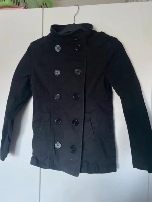 Svart skepparkavaj från H&M - Stilren svart skepparkavaj från H&M med knäppning. Jackan har två fickor framtill och klassisk krage. Barnstorlek 146🤍
