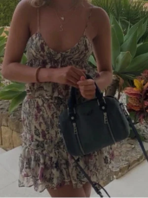 Blommig kort sommarklänning med volang - Söt kort kjol  med blommigt mönster i lila, rosa och beige toner. Klänningen har tunna axelband, v-ringning och volangdetaljer nertill. Perfekt för varma dagar och har en lätt och luftig känsla.