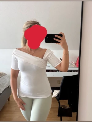 Vit asymmetrisk topp med offshoulder - Snygg vit topp med omlott-detalj och offshoulder-design. Toppen har korta ärmar och sitter figurnära. Perfekt för dig som gillar en stilren och trendig look.