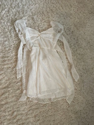 Vit kortklänning med puffärmar - Söt vit kortklänning med transparenta puffärmar och knytband. Klänningen har ett diskret prickigt mönster och omlottdetalj vid bysten. 