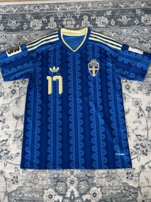 Blå Sverige fotbollströja nr 17 - Säljer en blå fotbollströja från Sveriges landslag med nummer 17 och namnet Gyökeres på ryggen. Tröjan har gula detaljer, mönstrad design och broderat landslagsemblem på bröstet. Perfekt för dig som vill stötta Sverige på match!