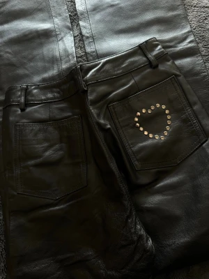 Svarta skinn/läderbyxor med nitar - Snygga svarta skinn/läderbyxor med raka ben och hög midja. På ena bakfickan finns ett hjärta av silverfärgade nitar som ger en cool detalj. Byxorna har bälteshällor och klassiska fickor både fram och bak. Tyvärr lite oklart vilken strl det är exakt men skulle gissa på XXS/XS.