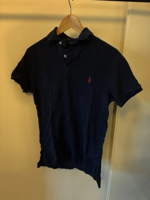 Polo Raulph Lauren pikétröja - Snygg marinblå pikétröja med korta ärmar och klassisk krage. Tröjan har två knappar vid halsen och en liten röd broderad logga på bröstet. Perfekt till jeans eller shorts för en clean look.