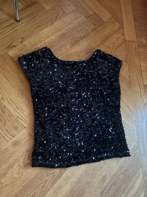 Svart paljettopp med korta ärmar - Säljer en svart topp täckt med glittrande paljetter. Toppen har korta ärmar och en rak passform, perfekt för dig som vill sticka ut. Materialet känns mjukt och följsamt mot kroppen. Storlek 152