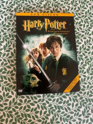Harry Potter och Hemligheternas kammare DVD - Säljer en DVD-utgåva av 'Harry Potter och Hemligheternas kammare' med två skivor. Omslaget är svart med färgstarka illustrationer av Harry, Hermione, Ron och husalfen Dobby. Perfekt för dig som samlar på Harry Potter eller vill se filmen hemma.