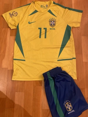 Brasilien fotbollströja och shorts Nike - Säljer ett Brasilien fotbollskit från Nike med gul tröja och blå shorts. Tröjan har gröna detaljer, nummer 11 och CBF-logga på bröstet. Shortsen är blå med grönt inslag, nummer 11 och Brasil-logga. Perfekt för match eller träning! Bra skick, finns ett litet hål som man ej knappt ser. Storlek 176