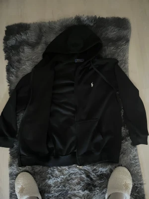 Svart hoodie från Polo Ralph Lauren - Snygg svart hoodie från Polo Ralph Lauren med dragkedja framtill och klassisk huva. Ikoniska vita loggan broderad på bröstet. Mjuk bomullskänsla och två fickor framtill. Perfekt för en chill och stilren look.