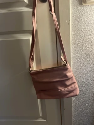 Ljusrosa axelväska i konstläder - Söt ljusrosa axelväska med justerbar rem och dragkedja upptill. Väskan har en mjuk, lite rundad form och flera fack inuti för bra organisering. Materialet är slätt konstläder och insidan är ljus med extra fickor. Perfekt för dig som vill ha en stilren och enkel väska.