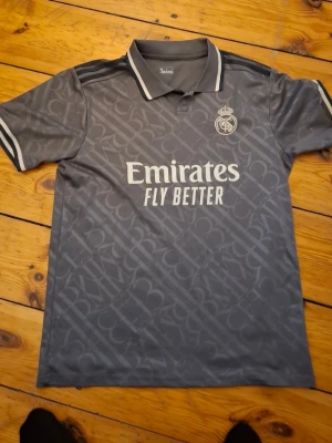 Grå Real Madrid matchtröja - Snygg grå Real Madrid matchtröja med vita detaljer och klubbmärke på bröstet. Tryck med 'Emirates Fly Better' på framsidan och 'Vini Jr. 7' på ryggen. Tröjan har krage och korta ärmar, perfekt för fotboll eller träning.