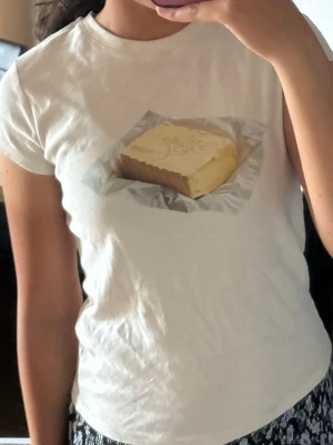 Vit t-shirt med smörtryck - Vit t-shirt med ett tryck av ett smörpaket på bröstet. Klassisk rund hals och korta ärmar. Tillverkad i mjukt bomullsmaterial för skön känsla. 