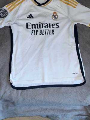 Real Madrid matchtröja Adidas vit - Säljer en vit Real Madrid matchtröja från Adidas med svarta och guldiga detaljer. Tröjan har klubbmärke, Adidas-logga och Emirates Fly Better-tryck på bröstet samt guldiga ränder på axlarna. Tillverkad i lätt och ventilerande material.