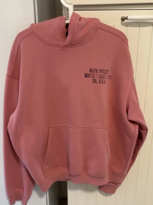 Flamingorosa hoodie  - En flamingorosa hoodie med Napa Valley tryck i Storlek L i lite oversize passform i armar och kortare längd så kan fungera som en Medium också.